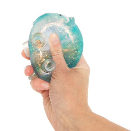 Hand squeezing a blue glitter squishy pig toy, showing the legs popping out and glitter shifting inside