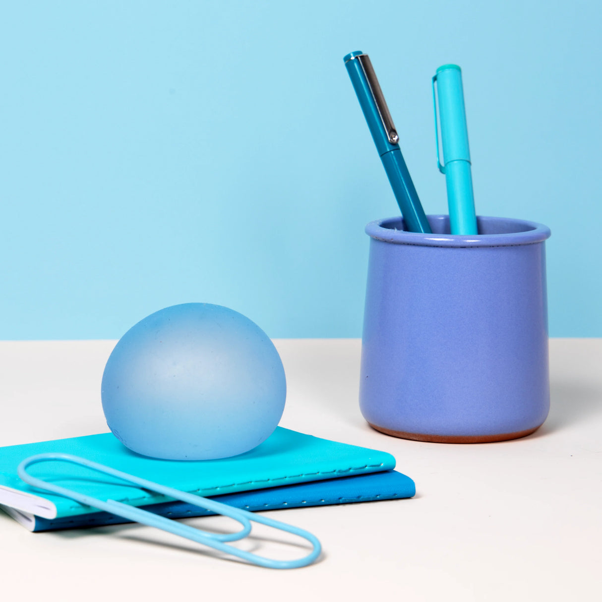 Pale blue Dew Speks Gump Original stress ball on a teal notebook beside a pen pot and paperclip