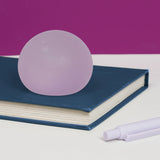 Lilac Mist Speks Gump Original stress ball on a dark blue book beside a pen against a purple background