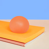 Coral orange Reef Speks Gump Original stress ball resting on a yellow notebook against a blue background