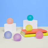 Six Speks Mini Gump stress balls in pastel shades displayed on white plinths and books against a blue background