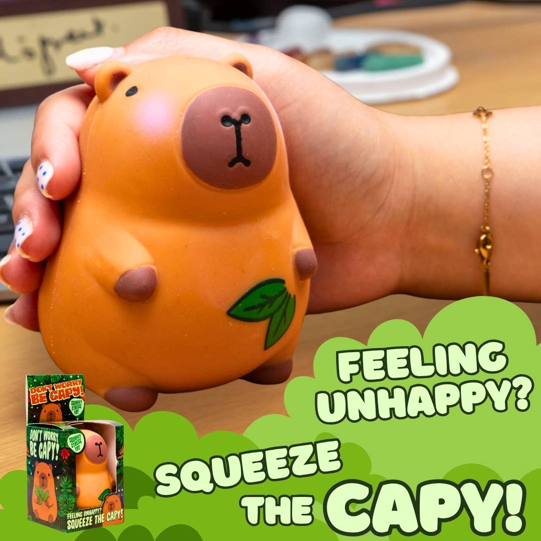 A hand holding a squeeze toy shaped like a capybara in orange, with a green leaf detail and the text 'FEELING UNHAPPY? SQUEEZE THE CAPY!' on a green graphic overlay, plus a small product box shown in the lower left.