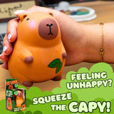 A hand holding a squeeze toy shaped like a capybara in orange, with a green leaf detail and the text 'FEELING UNHAPPY? SQUEEZE THE CAPY!' on a green graphic overlay, plus a small product box shown in the lower left.