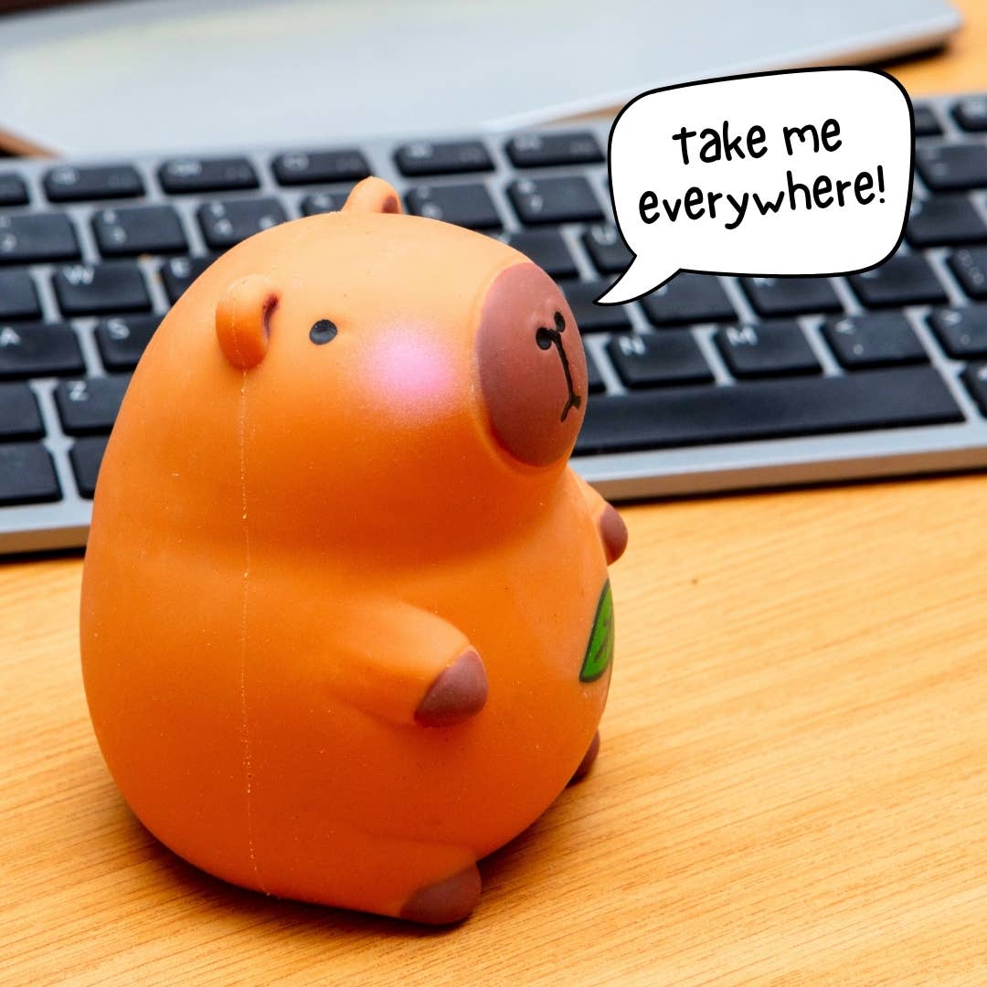 An orange pig-shaped squishy stress toy sitting on a wooden desk in front of a computer keyboard, with a speech bubble that says 'take me everywhere!'.