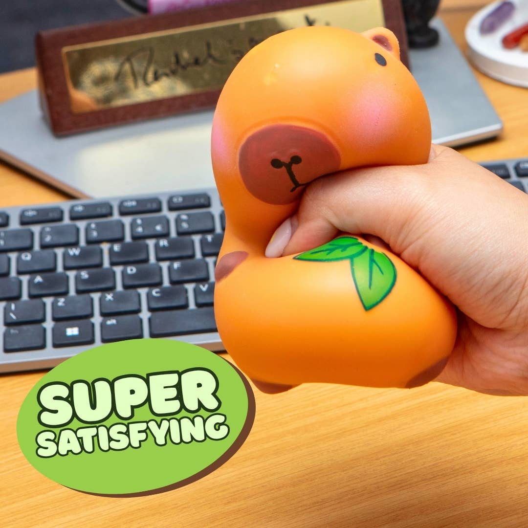 A hand squeezing an orange-colored squishy toy shaped like a duckling with a green leaf detail, set on a wooden desk in front of a keyboard. A green circular sticker overlay reads 'SUPER SATISFYING'.