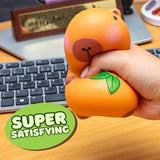 A hand squeezing an orange-colored squishy toy shaped like a duckling with a green leaf detail, set on a wooden desk in front of a keyboard. A green circular sticker overlay reads 'SUPER SATISFYING'.