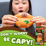 A woman holds an orange capybara-shaped squishy toy with a green leaf detail. The image includes a product box on the right and a large green promotional banner at the bottom that reads 'DON'T WORRY BE CAPY!'.