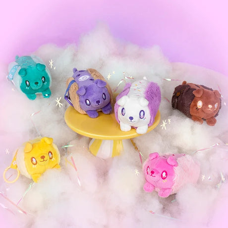 Six small plush animal toys in different colors are arranged on white fluffy material with a pastel pink background. The toys resemble cute animal characters and include teal, purple, white, brown, yellow, and pink versions.