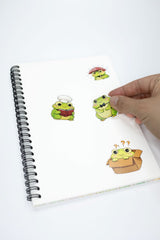 A hand is holding a sticker from a white spiral notebook. The notebook has three cartoon frog stickers on it, each with a different expression and outfit.