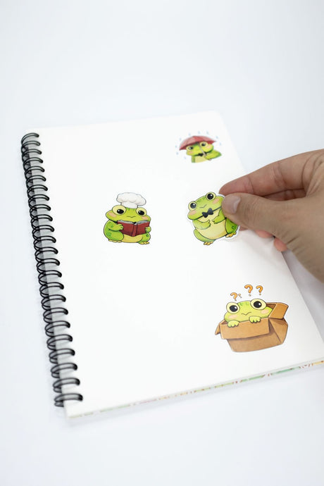 A hand is holding a sticker from a white spiral notebook. The notebook has three cartoon frog stickers on it, each with a different expression and outfit.
