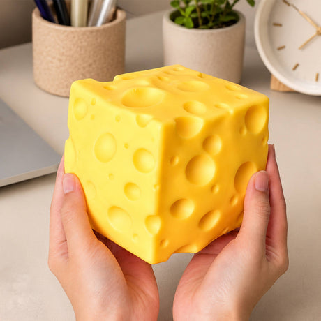 A person holds a yellow cheese-shaped squishy with realistic holes, shown in a home office setting with a pen cup, plant, and clock in the background.