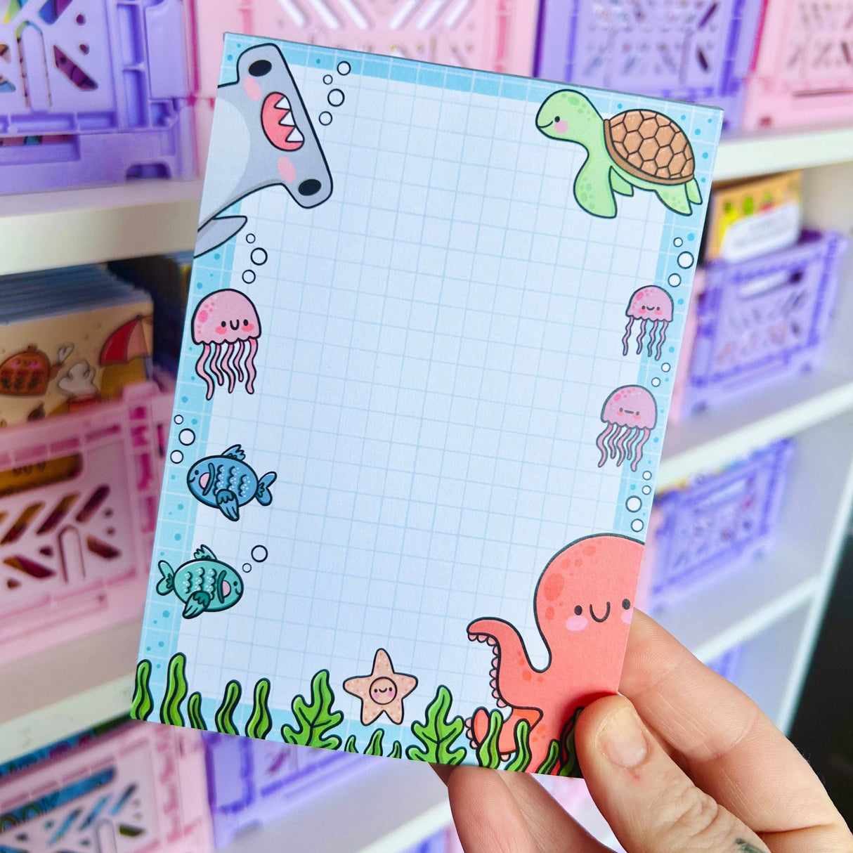 Adult hand holding the Under the Sea kawaii notepad in front of pastel storage shelves showing full ocean border design